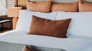 Ein ordentlich bezogenes Bett mit mehreren Kissen, einer Lampe und Blick ins Freie.