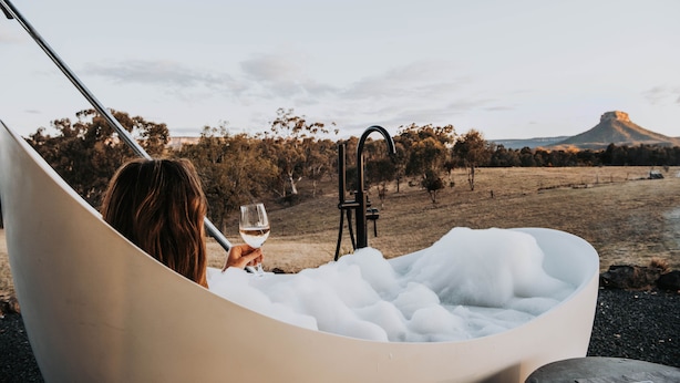 Eine Person entspannt sich in einer mit Schaum gefüllten Badewanne, mit Blick auf eine Landschaft und einen Berg im Hintergrund.