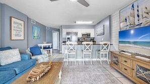 A modern living room with a blue sofa, a wooden coffee table, and a TV on a wooden stand. There is a kitchen area with white cabinets and a bar with white chairs.