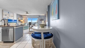 A compact kitchen with a refrigerator, a white island with blue cushioned stools, and a dining area with a view to the balcony.