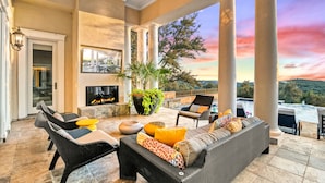 A covered outdoor seating area with wicker furniture, a fireplace, and a view of the water. - Fortaleza | Hill Country Estate with Infinity Pool, Tennis Court and 3 Acre Grounds (San Antonio)