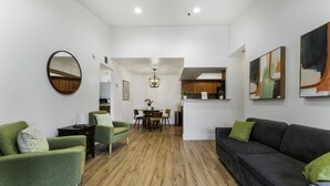 Een moderne woonkamer met een eetgedeelte, houten vloeren, groene fauteuils, een zwarte bank en abstracte wandkunst.
