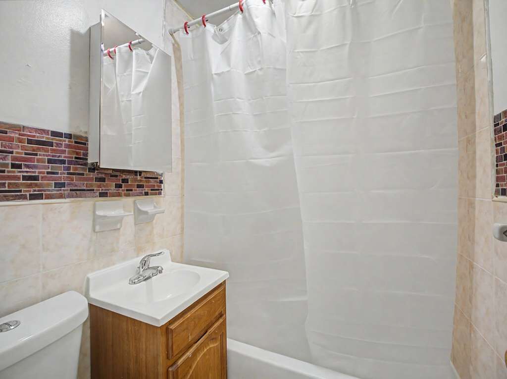 Un bagno con un lavandino bianco, un mobile bagno in legno, uno specchio, una tenda per la doccia e una parete rivestita di piastrelle con motivo a mattoni.