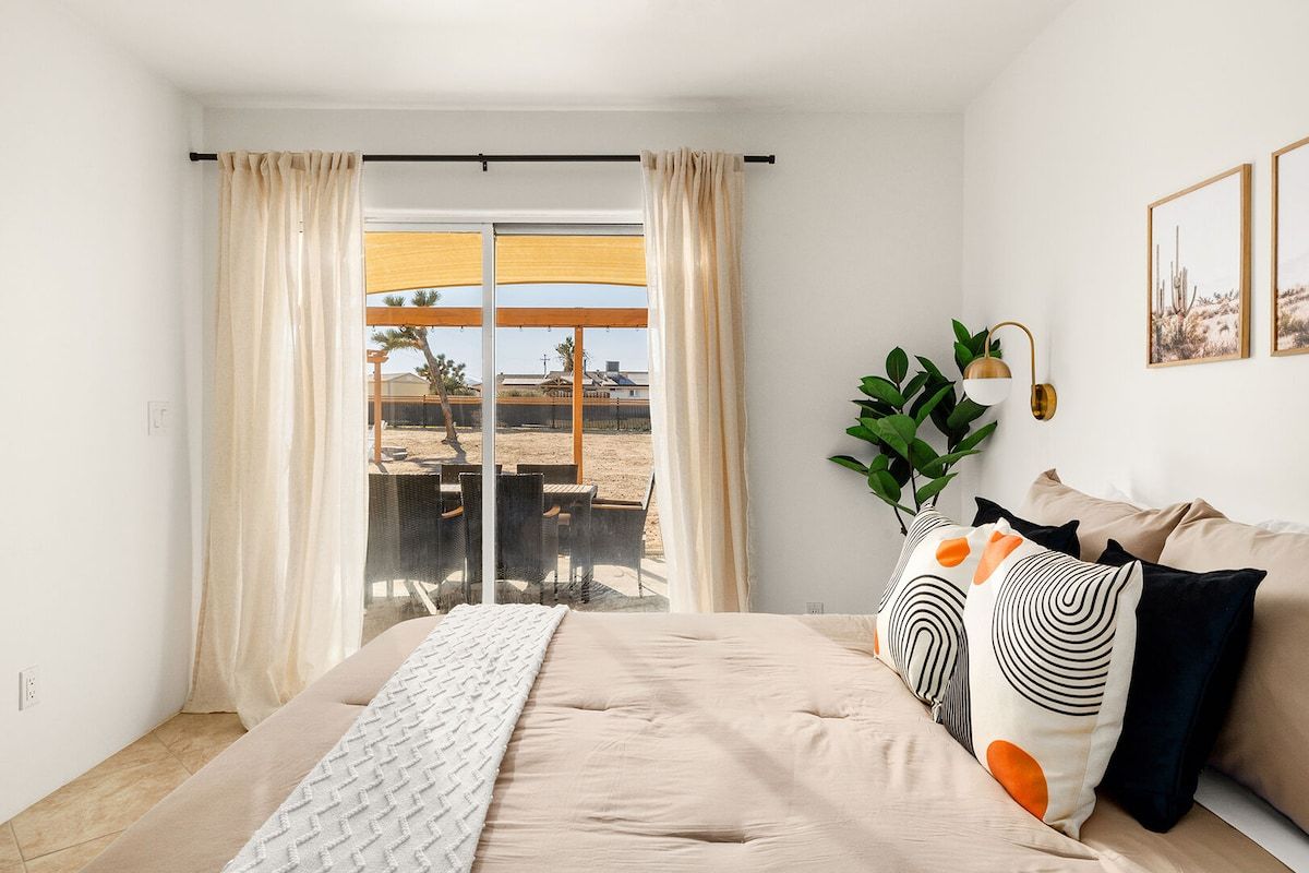 A bedroom with a bed, curtains, a view of the outside, a plant, and a wall-mounted lamp.