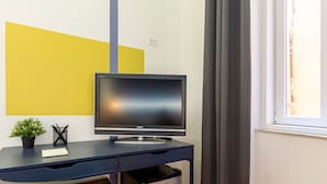 A modern room with a desk, a television, a trash bin, a potted plant, and a window with curtains. - Ghiberti - Lovely Guesta Room w Bathroom (Trieste)