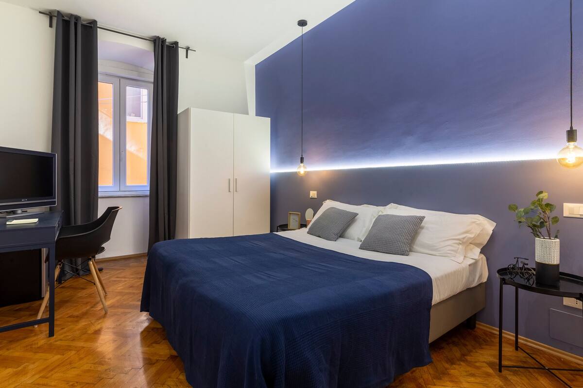 Un dormitorio con cama, escritorio, silla, armario y mesita de noche con lámpara.
