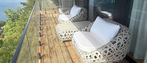 A balcony with wooden flooring and wicker furniture, overlooking a body of water and mountains.