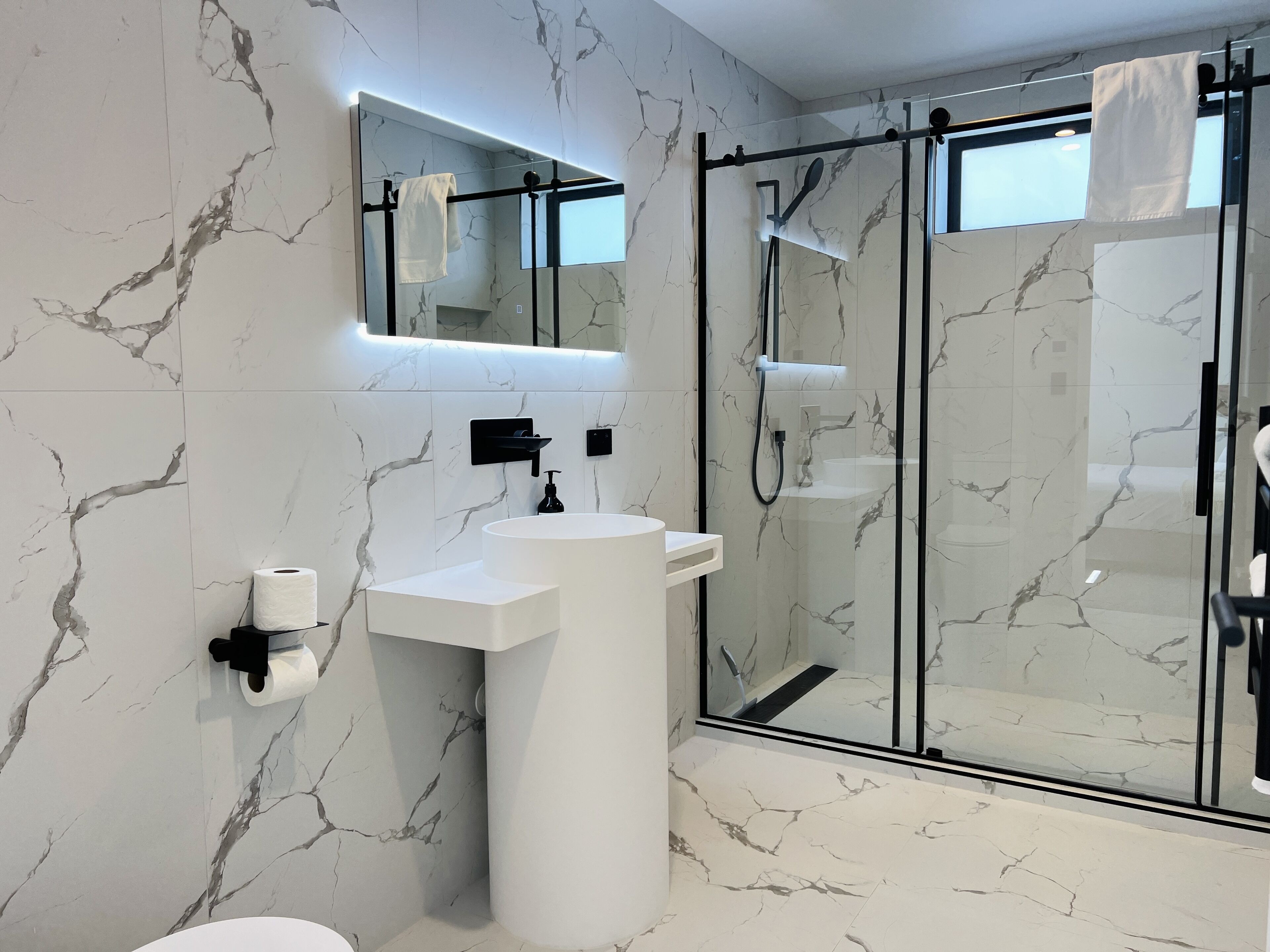 Ein modernes Badezimmer mit Marmor-Spritzschutz, einem weißen Waschbecken, einer Duschkabine aus Glas und einem Spiegel über dem Waschbecken.