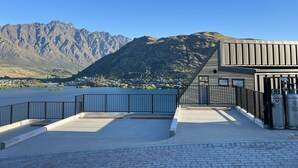 A waterfront building with a metal railing, a paved area, and a mountain backdrop.
