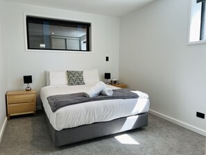 Une chambre moderne avec un grand lit, deux tables de chevet, une fenêtre et une tête de lit intégrée.