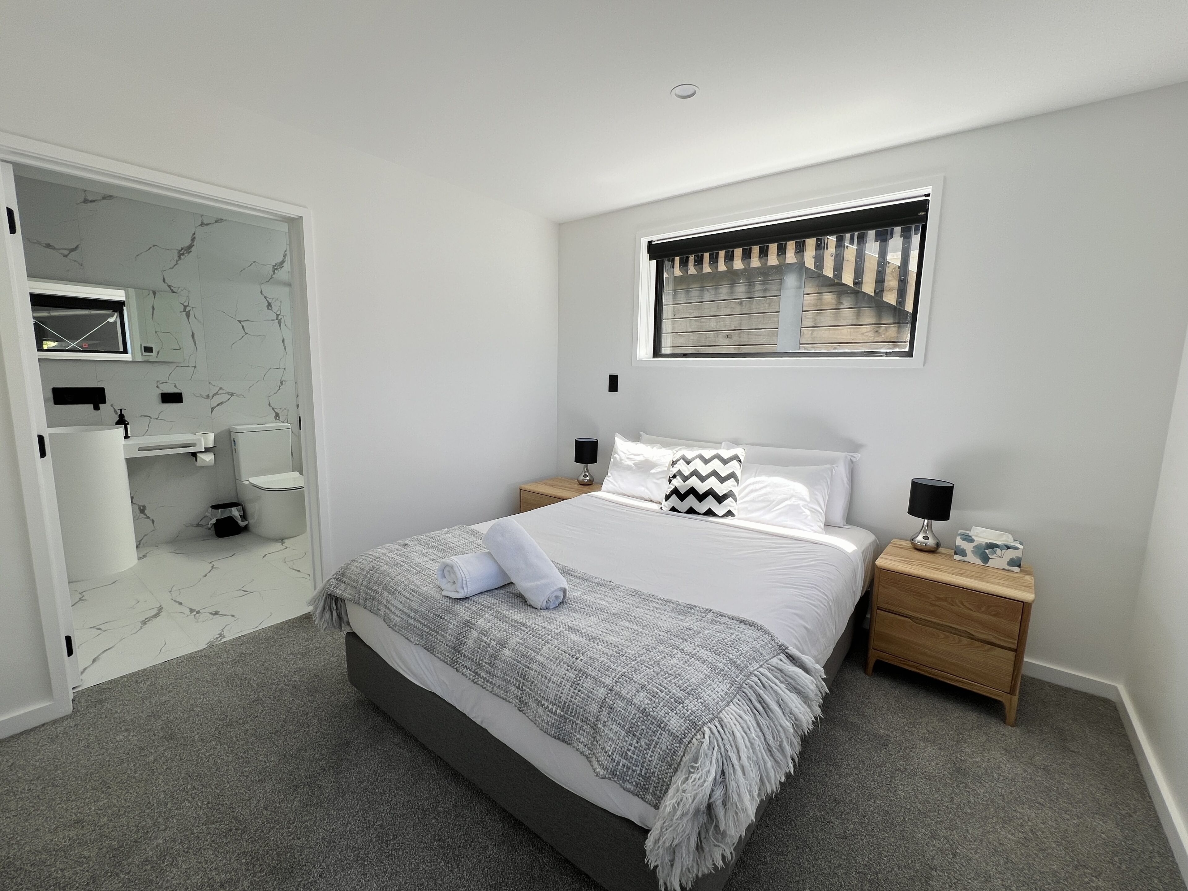 A modern bedroom with a bed, bedside tables, a window, and a bathroom with a toilet and sink.