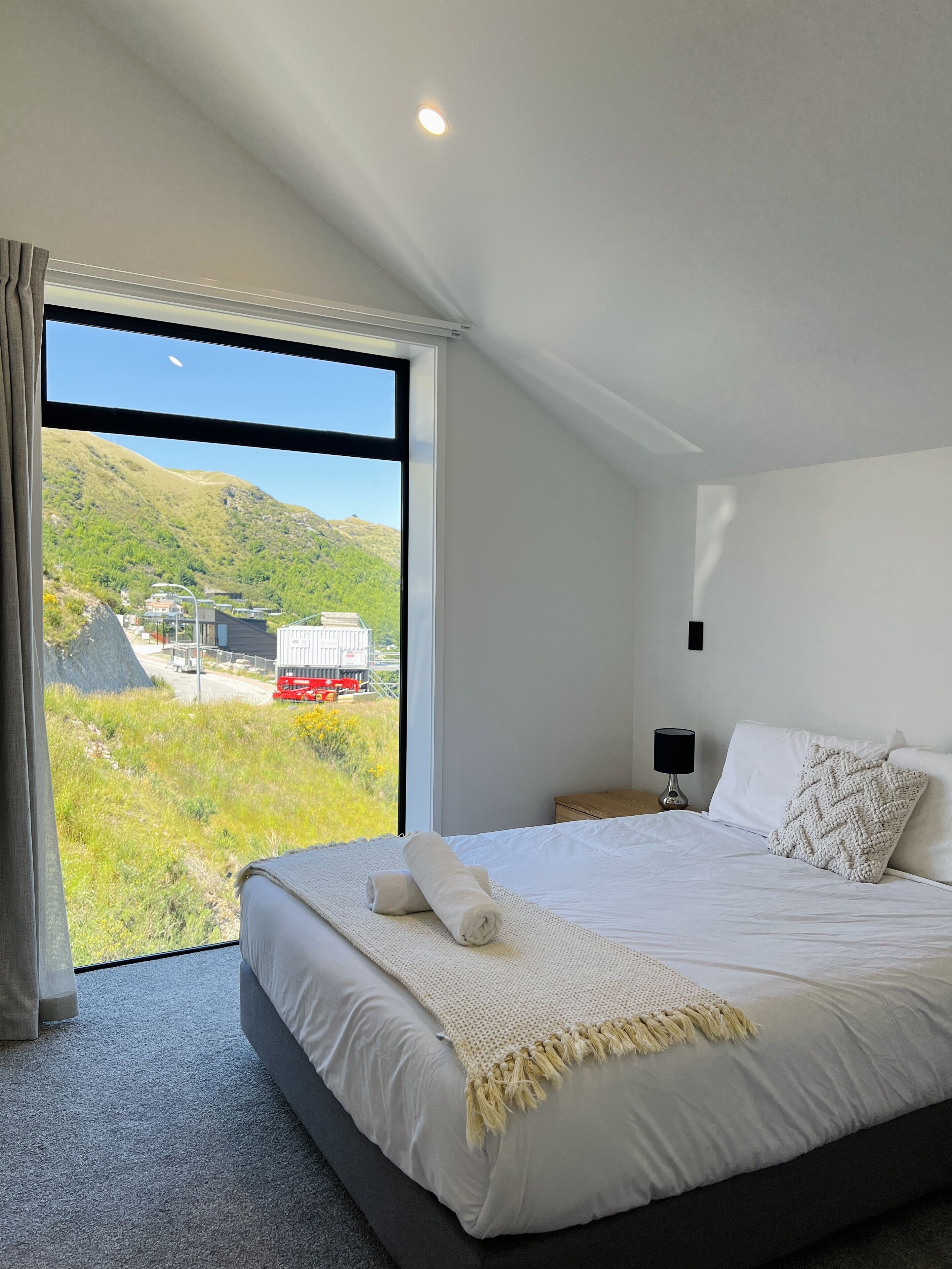 Una camera da letto con una grande finestra, un letto con lenzuola bianche, un comodino e vista su un paesaggio collinare con una strada.