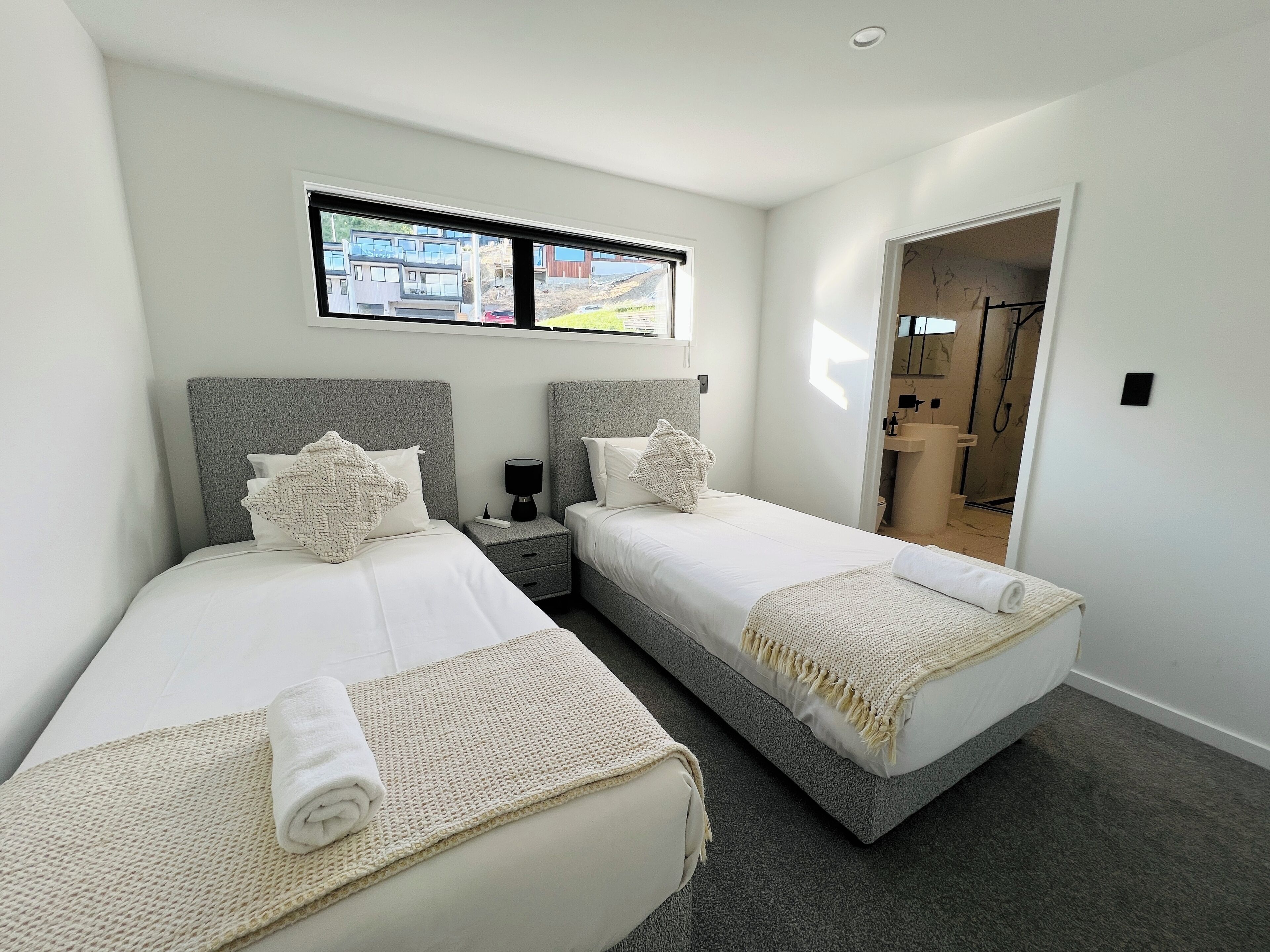A hotel room with two beds, a window with a view of buildings, and a bathroom visible through an open door.