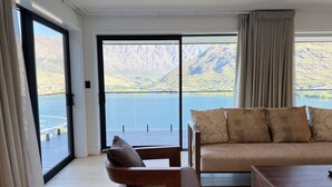 A modern living room with a sofa and a chair, large windows with a mountain view, and a wooden floor.