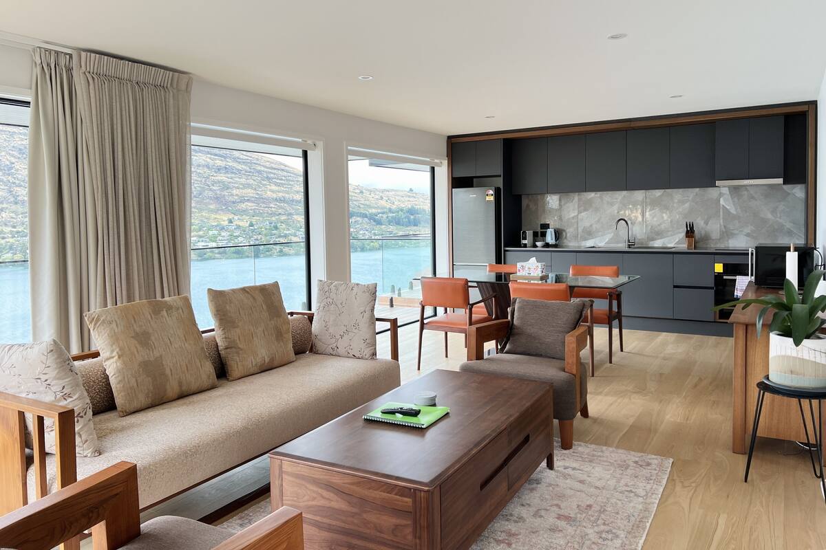 A modern living room with a sofa, coffee table, and dining area. A kitchen with dark cabinets and a view of the lake is visible.