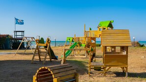 Une aire de jeux sur la plage, avec des balançoires, des toboggans et une cabane en bois.