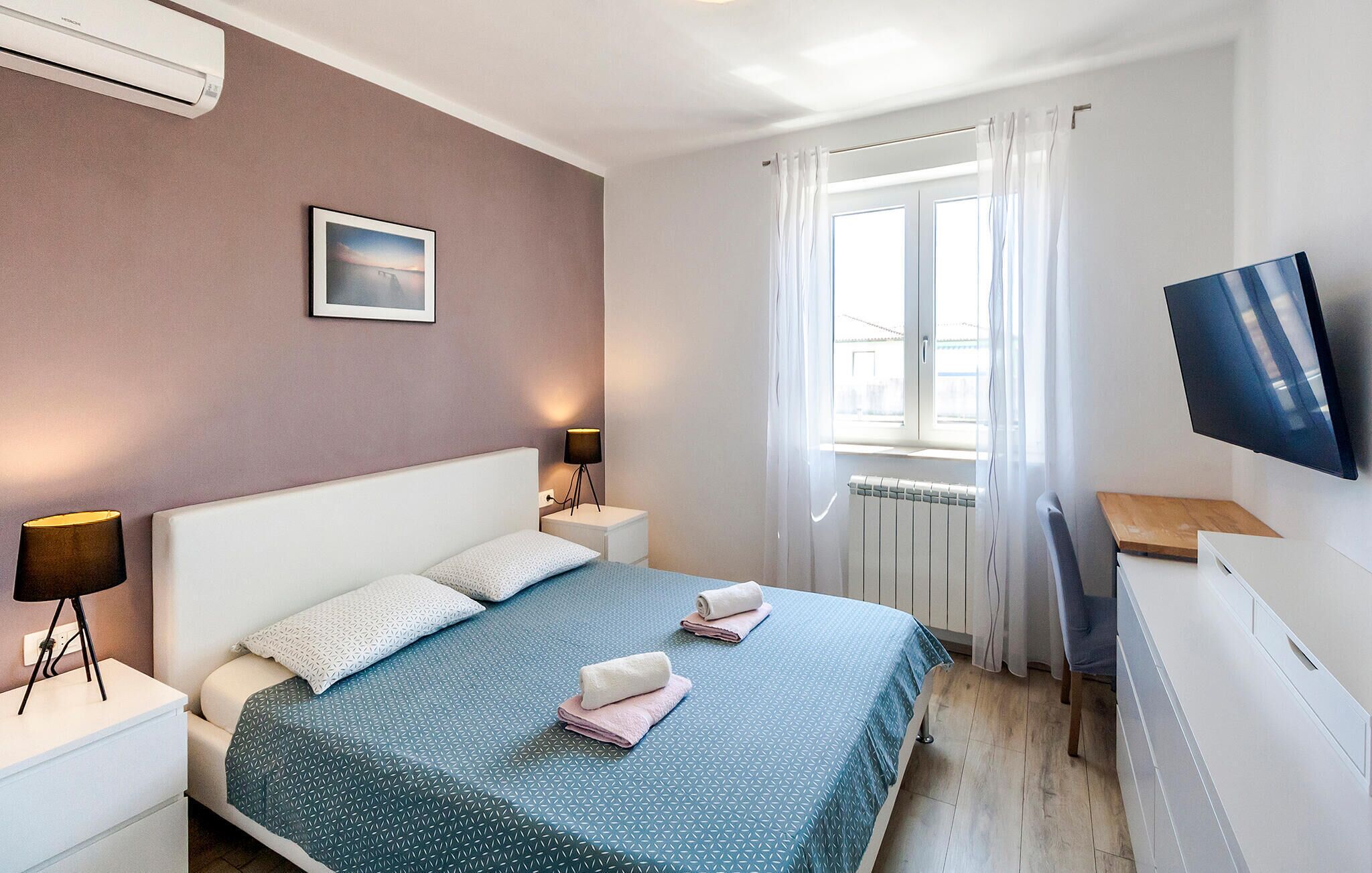 Une chambre d’hôtel avec un lit, une télévision, une fenêtre avec des rideaux, une table de chevet avec une lampe et un tableau au mur.