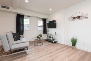 Ein modernes Wohnzimmer mit grauem Sofa, einem Glastisch, einem Flachbildfernseher und einem an der Wand befestigten Schild mit der Aufschrift "HAPPILY Ever After".