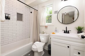 A bathroom with a white brick wall, a shower with a beige curtain, a toilet, a sink with a mirror, and a trash can.