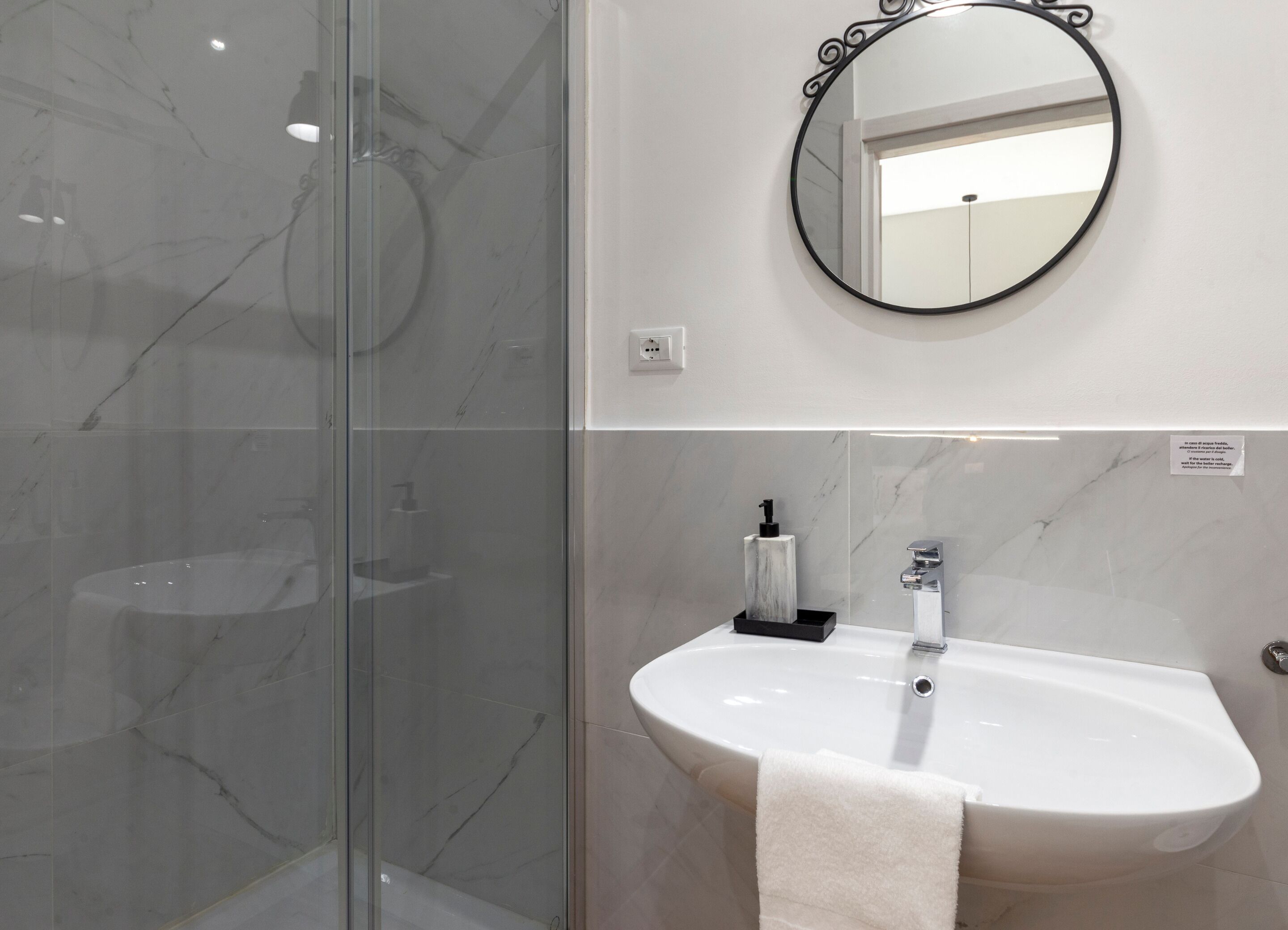 A modern bathroom with a glass shower enclosure, a round mirror, and a white sink with a towel.