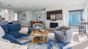 Een moderne woonkamer met een blauwe bank, grijze fauteuils, een salontafel en een open haard.