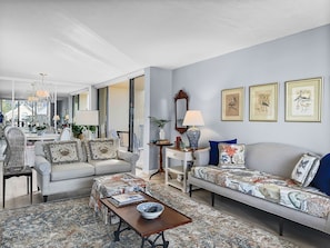 A living room with a sofa, armchairs, a coffee table, and a patterned rug. There is a dining area with a table and chairs visible through an open door.