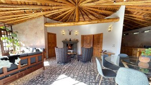 A spacious room with a stone-tiled floor, wooden ceiling beams, a fireplace, and a dining area with a glass table and chairs. - Cabañas Real Victoria (El Oro de Hidalgo)