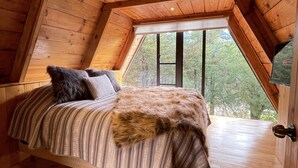 A loft bedroom with a sloped ceiling, wooden walls, and a large window. - Cabañas Real Victoria (El Oro de Hidalgo)