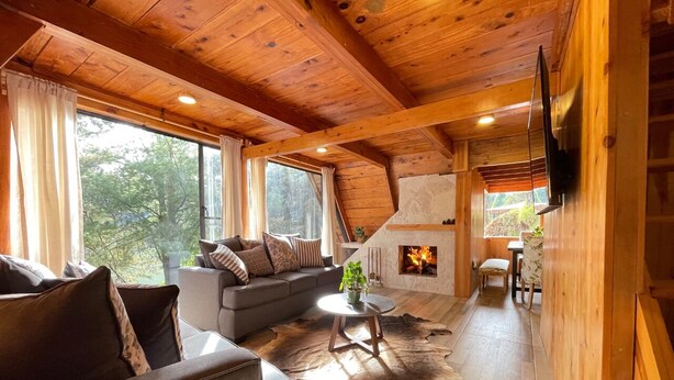 A cozy cabin interior with a wooden ceiling, a fireplace, a sofa, a coffee table, and a dining area. - Cabañas Real Victoria (El Oro de Hidalgo)