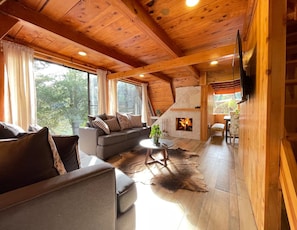 A cozy cabin interior with a wooden ceiling, a fireplace, a sofa, a coffee table, and a dining area. - Cabañas Real Victoria (El Oro de Hidalgo)