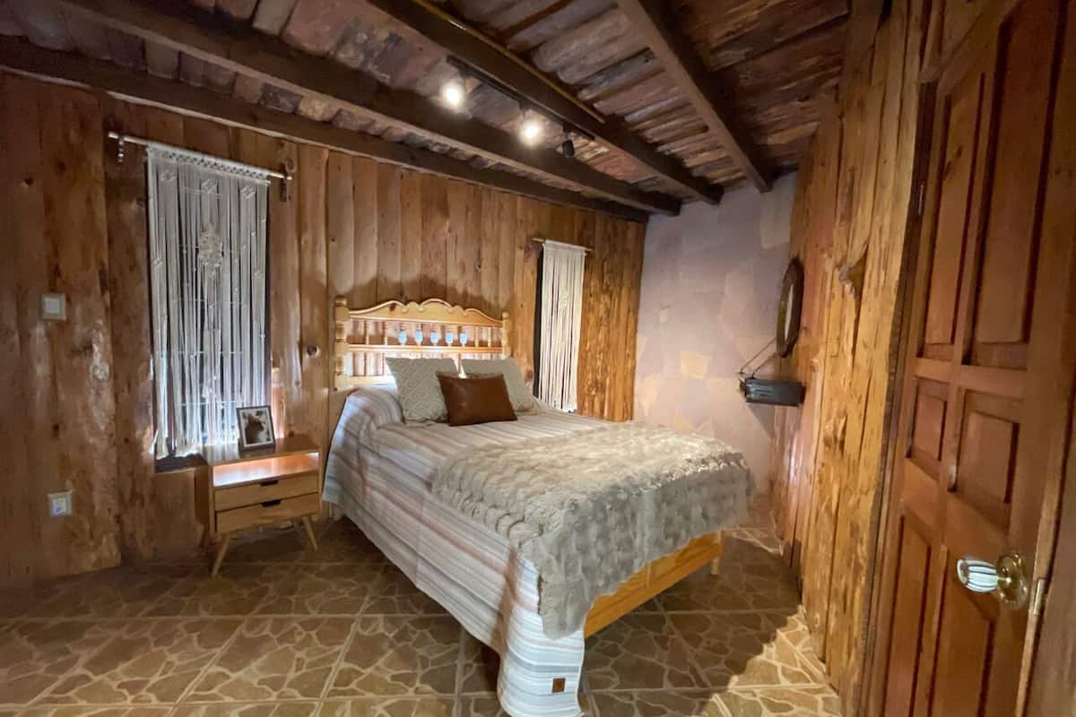 A wooden room with a bed, a nightstand, and a window with curtains.