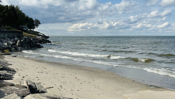 A beach with large rocks in the foreground, a sandy shore, and a view of the ocean under a partly cloudy sky.