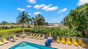 A poolside area with sun loungers and umbrellas, surrounded by palm trees and a view of the beach.