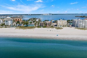 A coastal cityscape with a wide sandy beach, a marina with boats, and a variety of buildings including hotels and residential structures. - Gulf Beach 201 - Gulf-Front Serenity with Sunset Views (St Pete Beach)