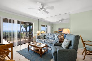 A spacious living room with a large sofa, a coffee table, and a chair. There is a balcony with a view of the ocean.