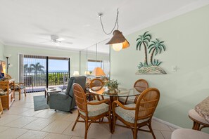 Een eetgedeelte met een ronde glazen tafel, rieten stoelen en uitzicht op de oceaan door grote schuifdeuren.