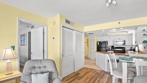 A modern kitchen with white cabinets, a microwave, and a coffee maker. There is a dining area with a glass table and white chairs.