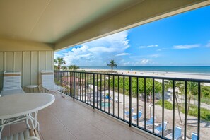 A balcony with a view of a beach, a pool, and outdoor seating.