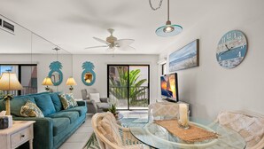 Ein modernes Wohnzimmer mit einem türkisfarbenen Sofa, Korbstühlen, einem Glastisch und einem Deckenventilator.