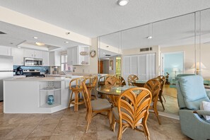 A spacious living area with a dining table surrounded by wicker chairs, a kitchen with modern appliances, and a large window providing natural light. - 🌊 Panoramic Gulf Views & Soothing Coastal Vibes (St Pete Beach)