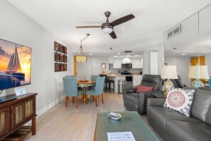 A modern living room with a flat-screen TV, dining area, kitchen, and a ceiling fan.