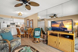 A living room with a television, wicker furniture, ceiling fans, and a kitchen in the background.