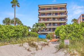 A multi-story building with balconies, surrounded by dense greenery and a sandy area.