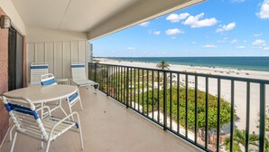 A balcony with a view of a beach, white chairs, and a table.