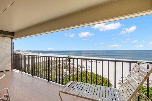 A balcony with a white metal chair, a view of the beach, and a clear sky.