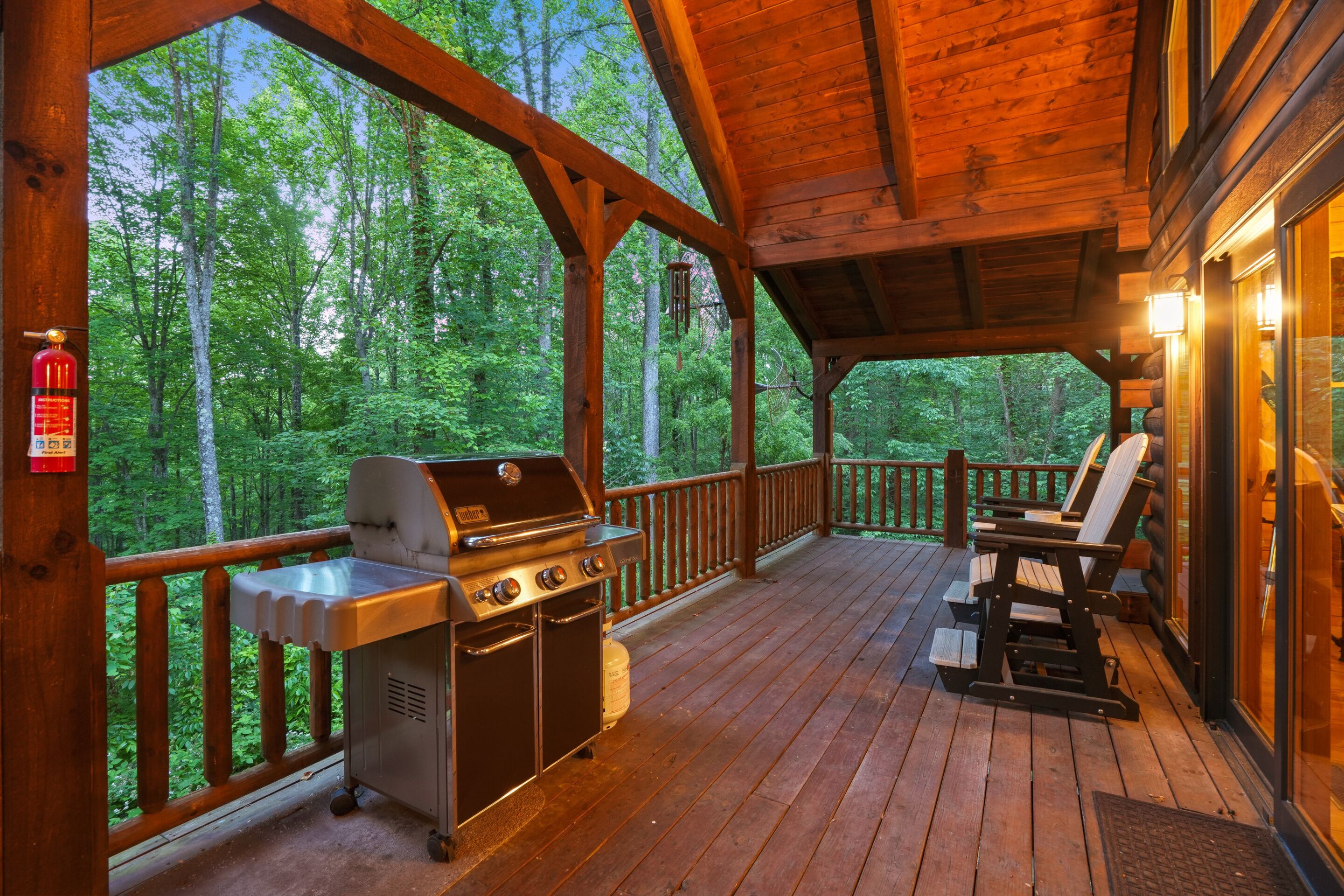 A covered outdoor deck with a grill and seating area, surrounded by a wooden railing and overlooking a forest.