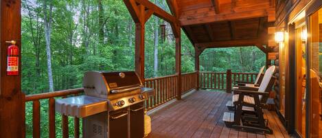 A covered outdoor deck with a grill and seating area, surrounded by a wooden railing and overlooking a forest.