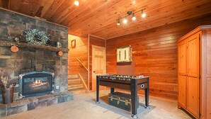 Una habitación acogedora con chimenea de piedra, paredes de madera, una mesa de fútbol y una puerta que da a otra zona.