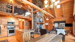 A rustic cabin interior with a stone fireplace, wooden dining table, and a kitchen with a stove and oven.