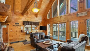 A cozy cabin interior with wooden walls, a fireplace, a dining area, and large windows offering views of the outdoors.